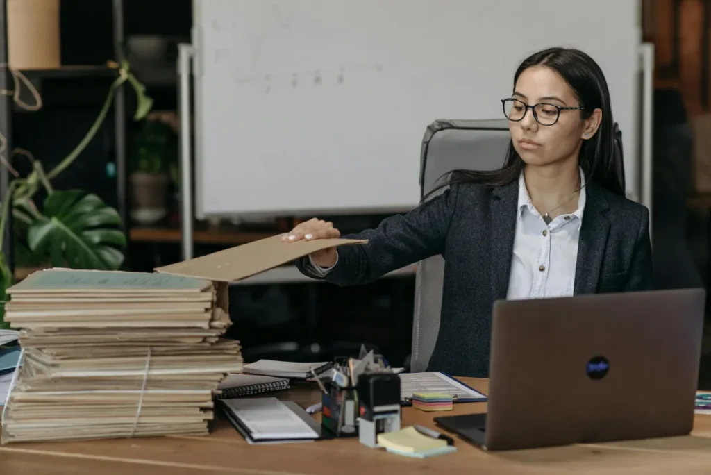 Seorang staf admin perempuan di kantor modern sedang mengorganisasi tumpukan dokumen di samping laptop